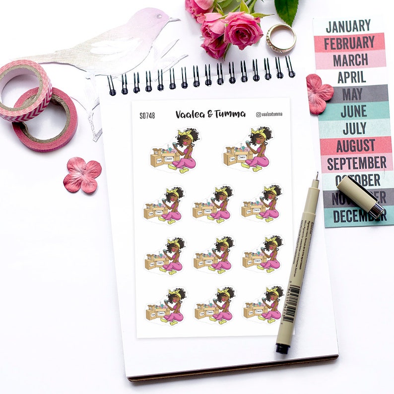 May include: A sheet of stickers featuring a cartoon illustration of a woman with brown skin, black hair, and a yellow headband. She is wearing a pink shirt and black pants and is holding a box with a yellow lid. The sticker sheet is labeled "Vaalea & Tumma" and has the number "S0748" printed on it.