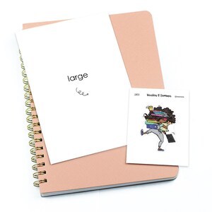 May include: A pink notebook with a gold spiral binding and a white cover sheet with the word "large" written on it. A sticker with a cartoon illustration of a woman carrying a stack of books is attached to the notebook.