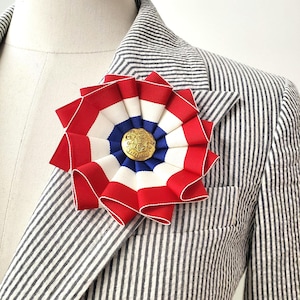 Könnte beinhalten: Eine patriotische Rosettennadel mit rot-weiß-blauem Band, in der Mitte ein goldfarbener Knopf. Die Nadel ist an einem schwarz-weiß gestreiften Blazer befestigt und sorgt für einen klassischen und stilvollen Look.