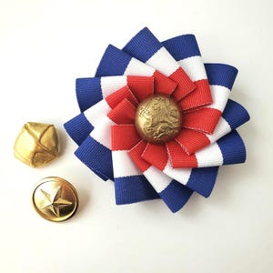 May include: A rosette badge featuring blue, white, and red striped ribbon arranged in a circular pattern with a central gold-toned button embossed with a lion and sword design, accompanied by two separate gold-toned buttons with star and geometric motifs.