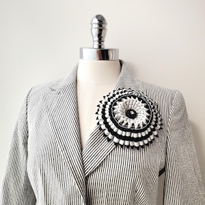 Könnte beinhalten: Eine schwarz-weiße Stoffblumenbrosche, die an einem gestreiften Blazer befestigt ist. Die Brosche hat Schichten aus schwarz-weißem plissiertem Stoff mit einem gepunkteten Mittelteil. Der Blazer ist schwarz-weiß gestreift.