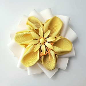 May include: A handmade flower hair accessory. The flower features layers of white and yellow fabric petals. The center of the flower has a yellow and orange decorative element. The accessory is designed to be worn in the hair.