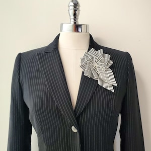 Op de afbeelding: Een zwarte blazer met krijtstreep, voorzien van een enkele witte knoopsluiting en een grote, decoratieve stoffen broche op de revers. De broche is gemaakt van geplooid zwart-wit gestreept textiel in een cirkelvormig waaierpatroon.