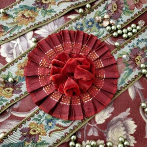 Könnte beinhalten: Nahaufnahme einer roten Rosette mit Samtmittelteil, umgeben von plissiertem Band. Die Rosette liegt auf einem Stoff mit Blumenmuster mit dekorativen Zierleisten und verstreuten Perlen.