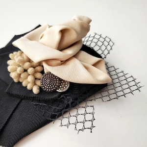 May include: A decorative hair accessory featuring a beige fabric bow, a cluster of small beige textured spheres, and two circular metallic buttons with a dark finish, all layered over a black ribbed fabric base with black netting accents.