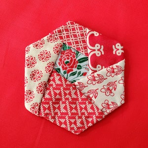 May include: A red, white, and green fabric coaster with a geometric pattern. The coaster is made of six pieces of fabric sewn together in a star shape.