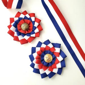 May include: Two circular rosettes made of red, white, and blue striped grosgrain ribbon. One rosette features a brass button with an embossed lion emblem, while the other has a brass button with a five-pointed star. A long matching ribbon strip.