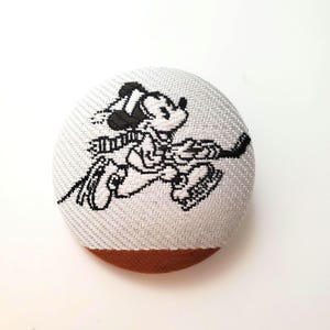 May include: A round, gray button with a brown bottom edge. The button features a black and white embroidered image of Mickey Mouse ice skating and holding a hockey stick. The button has a textured fabric appearance.