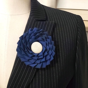 Könnte beinhalten: Eine marineblaue Stoffblumenbrosche mit weißer Mitte, befestigt an einem schwarzen Nadelstreifenblazer. Die Blume besteht aus geschichteten Bändern, die ein strukturiertes, kreisförmiges Design bilden. Der Blazer befindet sich auf einer Schaufensterpuppe.