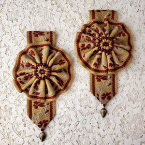 Peut inclure: Deux ornements décoratifs en tissu. Chacun présente un motif circulaire plissé doré avec des accents floraux rouges. Une bordure rouge encadre le cercle. Un ruban doré avec des motifs floraux rouges s'étend vers le bas, se terminant par un charme en forme de feuille.