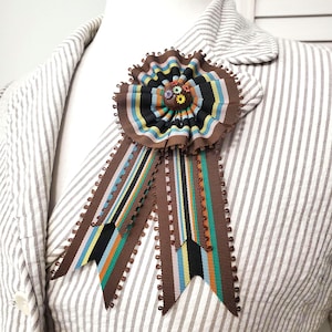 Könnte beinhalten: Eine braune Rosettenbrosche mit bunten Streifenbändern und einer kleinen Blumenverzierung. Die Brosche ist an einem gestreiften Blazer befestigt und bildet einen auffälligen Kontrast. Die Bänder haben einen gewellten Rand.