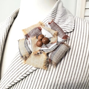 May include: A handmade fabric brooch with a flower design. The brooch features frayed edges and is made of tan, brown, and white striped fabric. Three brown buttons are in the center. The brooch is pinned to a striped blazer.