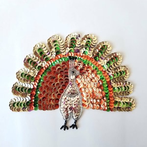 May include: A colorful peacock design made of sequins. The peacock is silver with a green, orange, and gold tail.