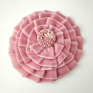 May include: A dusty pink velvet fabric flower brooch featuring multiple layers of pleated ribbon petals arranged in a circular pattern. A round, cream-colored button with an intricate, embossed floral and dot pattern sits at the center.