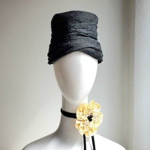 May include: A black fabric hat with a pleated design. The hat is on a white mannequin with a yellow flower brooch around the neck.