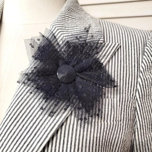 May include: A navy blue flower-shaped brooch made of layered tulle and a central button. The brooch is pinned to a striped blazer, creating a contrast of textures and patterns. The blazer has a classic seersucker stripe.