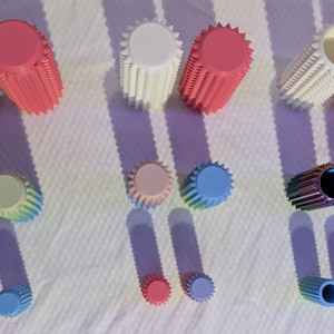 May include: An overhead shot displays several sets of colorful, cylindrical objects with a ridged design. The objects are in various colors, including white, pink, blue, and green. The objects are arranged on a white surface, casting long shadows.
