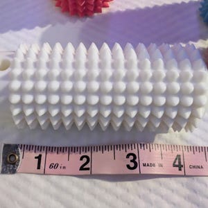May include: A white massage roller with a textured surface of rounded and pointed protrusions. The roller is approximately 5 inches long. Other similar rollers in pink and blue are in the background.