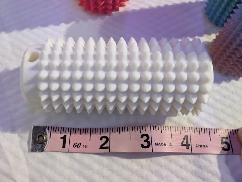 May include: A white massage roller with a textured surface of rounded and pointed protrusions. The roller is approximately 5 inches long. Other similar rollers in pink and blue are in the background.