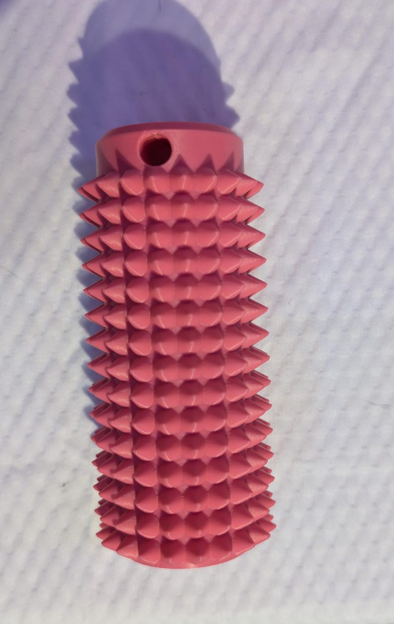 May include: A pink, cylindrical object with a series of conical protrusions. The top of the object has a small hole. The object is likely a massage tool or a similar device.