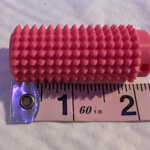 May include: A pink, cylindrical tool with a textured surface of evenly spaced, conical protrusions. The tool is approximately 2 inches long and is next to a pink measuring tape.