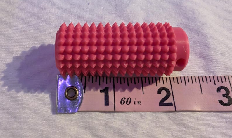 May include: A pink, cylindrical tool with a textured surface of evenly spaced, conical protrusions. The tool is approximately 2 inches long and is next to a pink measuring tape.