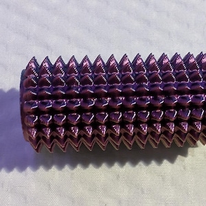 May include: A cylindrical, purple object with a series of triangular, spike-like protrusions. The object is metallic and reflects light, creating highlights. The item is set against a white background, with a shadow.