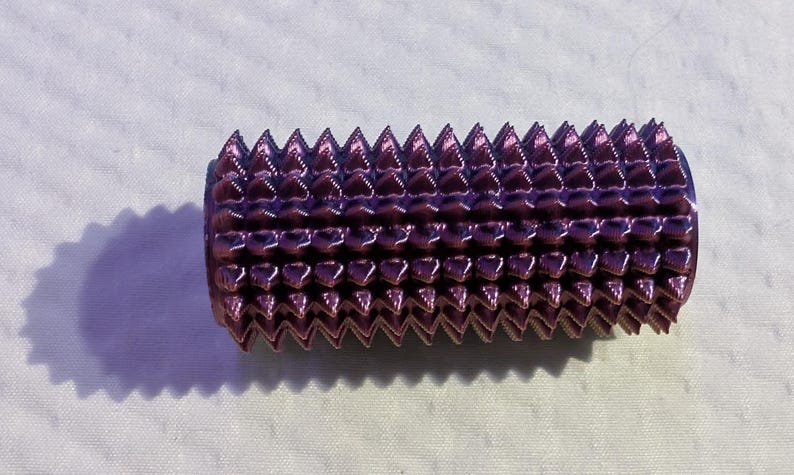 May include: A cylindrical, purple object with a series of triangular, spike-like protrusions. The object is metallic and reflects light, creating highlights. The item is set against a white background, with a shadow.