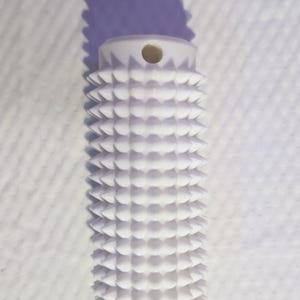 May include: A white, cylindrical object with a textured, spiky surface. The top has a small hole. The object casts a shadow on a white, textured background. The design is geometric and repetitive.