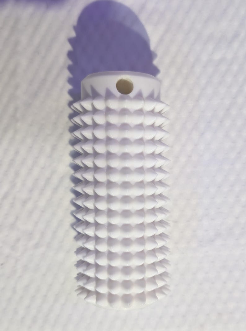 May include: A white, cylindrical object with a textured, spiky surface. The top has a small hole. The object casts a shadow on a white, textured background. The design is geometric and repetitive.