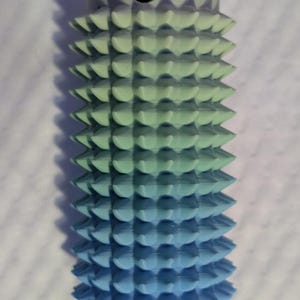 May include: A 3D-printed object with a gradient color scheme, transitioning from light green to blue. The cylindrical shape features a series of conical spikes. The top has a small hole. The object is likely a decorative or functional item.