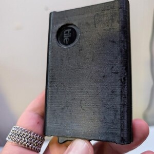 May include: A black rectangular Tile tracker with a rounded corner and the Tile logo. The tracker is held in a hand, and the surface has a slightly textured appearance. The tracker is designed to help locate items.
