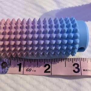 May include: A gradient-colored massage roller, transitioning from pink to blue, with a textured surface of conical spikes. The roller is approximately 3 inches long and is placed next to a pink measuring tape.