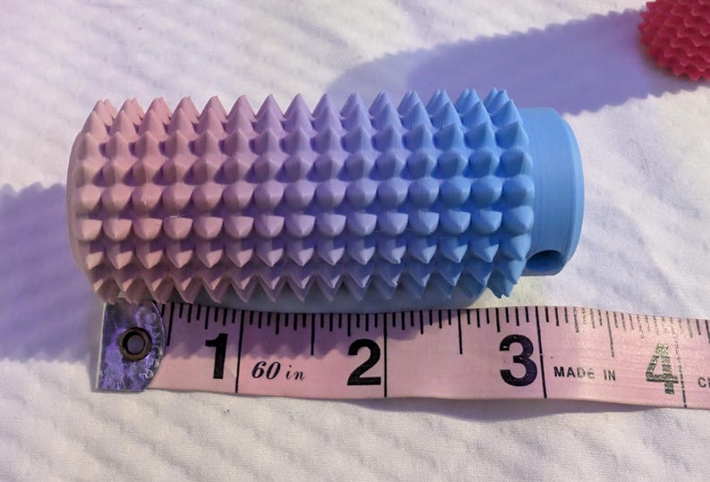 May include: A gradient-colored massage roller, transitioning from pink to blue, with a textured surface of conical spikes. The roller is approximately 3 inches long and is placed next to a pink measuring tape.