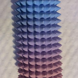 May include: A 3D-printed, gradient-colored object, transitioning from blue to purple to pink. The cylindrical form features a series of conical spikes. A hole is visible at the top. The object is likely a decorative or functional item.