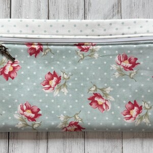 May include: A rectangular pouch with a floral pattern. The pouch features a light blue background with white polka dots and red and pink flowers. A zipper runs across the top, and the top edge is a light blue fabric with white polka dots.