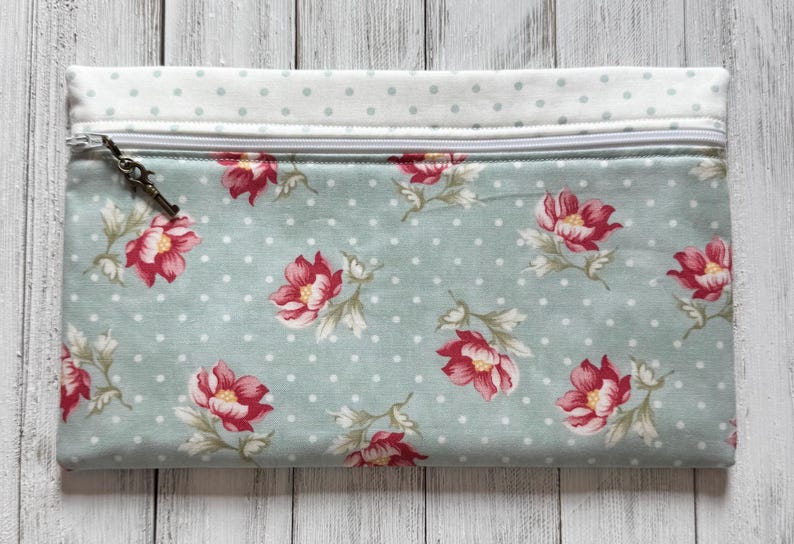 May include: A rectangular pouch with a floral pattern. The pouch features a light blue background with white polka dots and red and pink flowers. A zipper runs across the top, and the top edge is a light blue fabric with white polka dots.