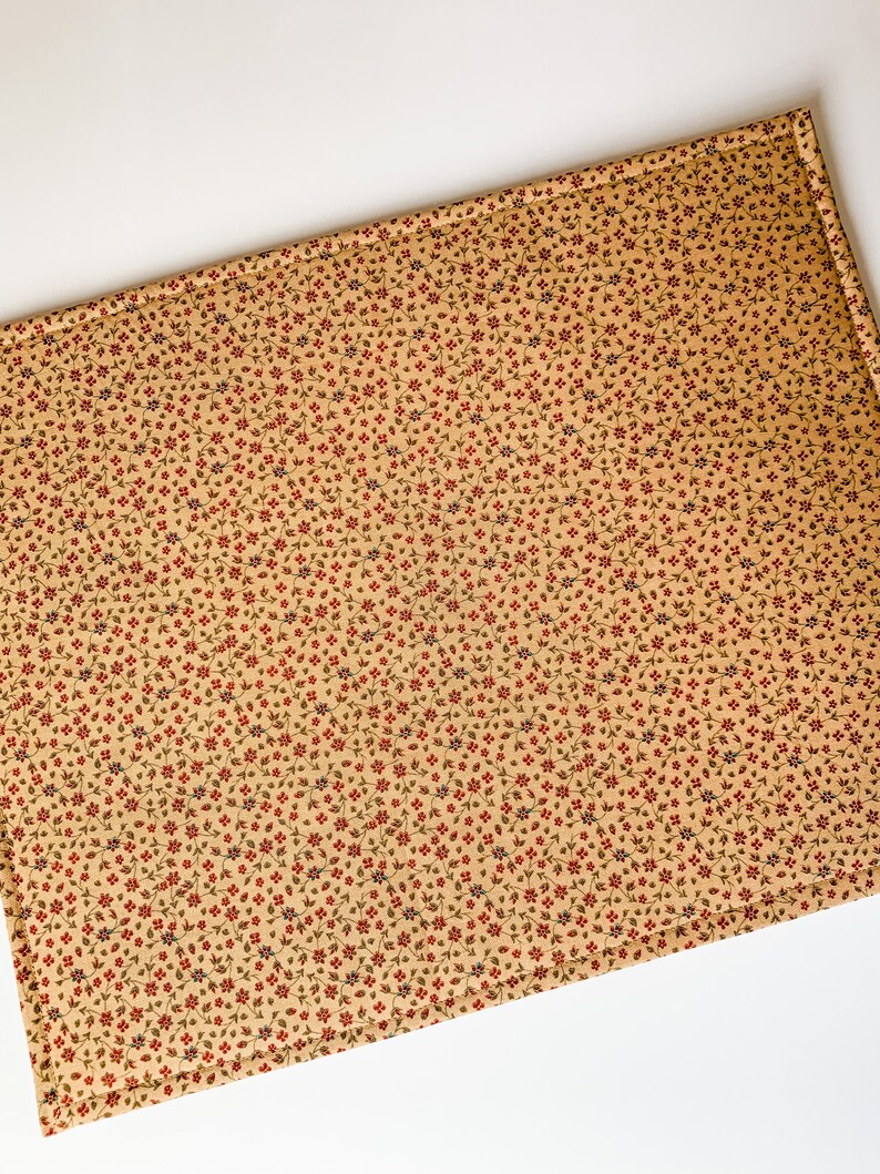 May include: A beige placemat with a small red and blue floral pattern. The placemat has a stitched border.