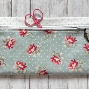 May include: A rectangular fabric pouch with a zipper closure, featuring a floral pattern of red flowers on a light blue background with white polka dots. A pair of red scissors is partially visible. The pouch has a white polka dot top.