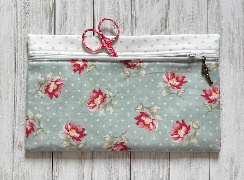 May include: A rectangular fabric pouch with a zipper closure, featuring a floral pattern of red flowers on a light blue background with white polka dots. A pair of red scissors is partially visible. The pouch has a white polka dot top.
