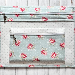 May include: A quilted organizer set featuring a large rectangular mat and a smaller zippered pouch. The set is light blue with a floral pattern of red flowers and white polka dots. The zippers have decorative key-shaped pulls.