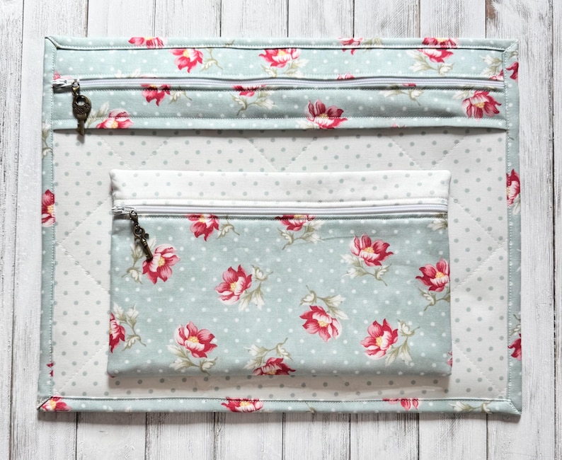May include: A quilted organizer set featuring a large rectangular mat and a smaller zippered pouch. The set is light blue with a floral pattern of red flowers and white polka dots. The zippers have decorative key-shaped pulls.