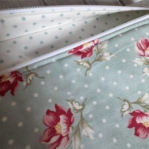 May include: A partially open, floral-patterned pencil case. The case features a light blue background with white polka dots and red floral designs. The interior lining is a lighter blue with small, evenly spaced dots. A white zipper runs along the top.