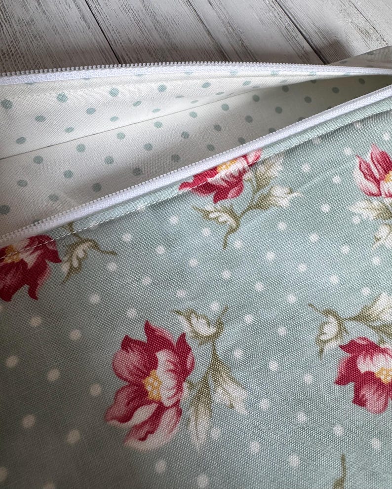 May include: A partially open, floral-patterned pencil case. The case features a light blue background with white polka dots and red floral designs. The interior lining is a lighter blue with small, evenly spaced dots. A white zipper runs along the top.