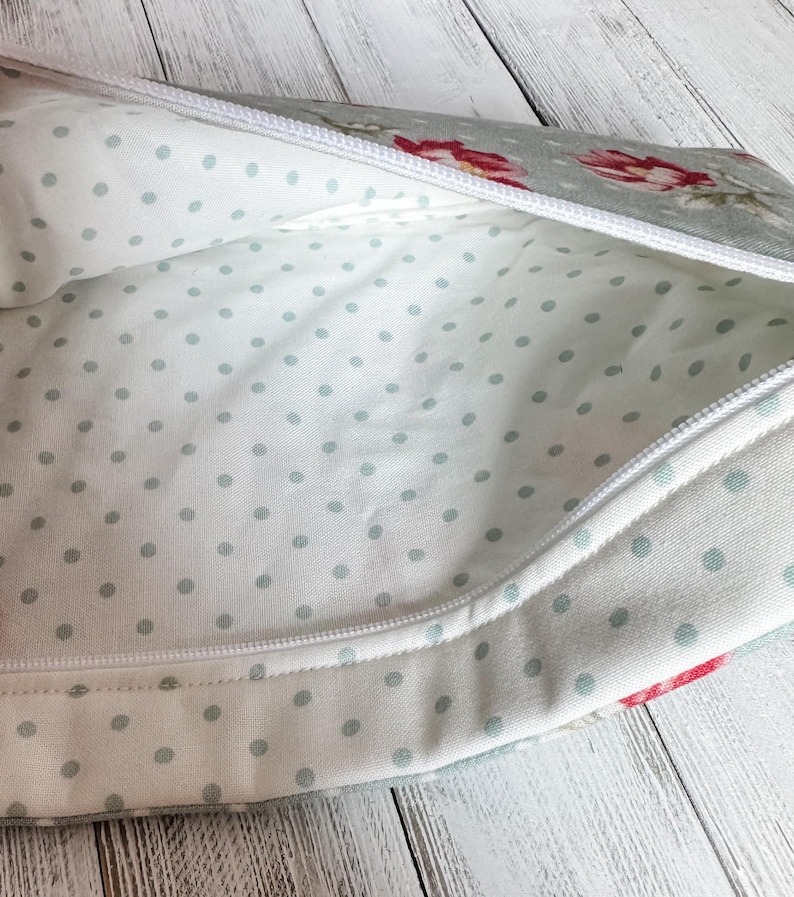 May include: Open view of a floral patterned zippered pouch. The exterior fabric is a light gray with red and pink flowers. The interior is white with small, light blue polka dots. A white zipper runs along the top.