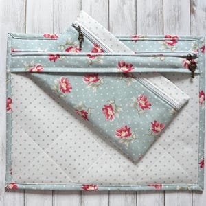 May include: A quilted, light blue fabric pouch with a floral pattern of red flowers and green leaves. A smaller, matching zippered pouch is inside. The larger pouch has a zipper with a key-shaped pull.