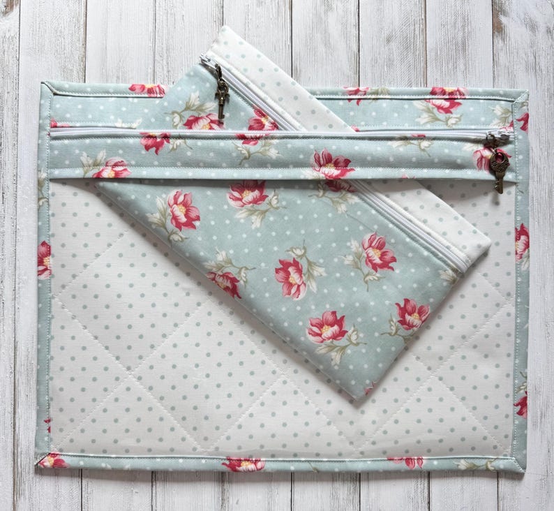 May include: A quilted, light blue fabric pouch with a floral pattern of red flowers and green leaves. A smaller, matching zippered pouch is inside. The larger pouch has a zipper with a key-shaped pull.