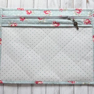 May include: A quilted fabric case with a floral pattern and a zipper closure. The case features a light blue background with red flowers and a key-shaped zipper pull. The interior is white with gray polka dots.