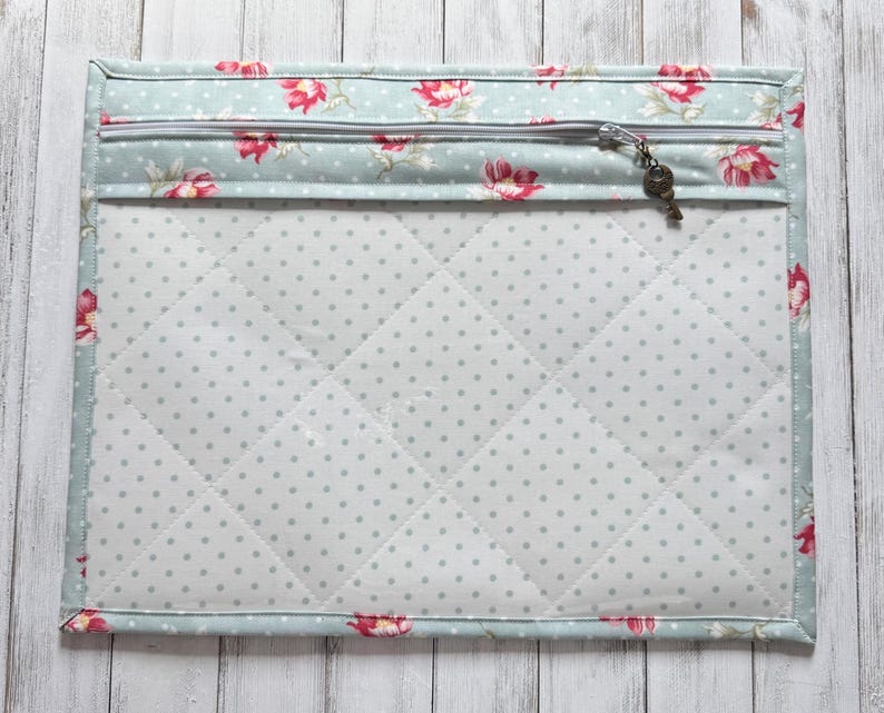 May include: A quilted fabric case with a floral pattern and a zipper closure. The case features a light blue background with red flowers and a key-shaped zipper pull. The interior is white with gray polka dots.