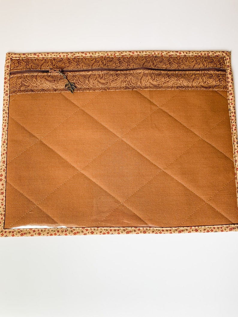 May include: A brown quilted fabric pouch with a zipper closure and a floral patterned trim. The pouch has a clear plastic window on the front.